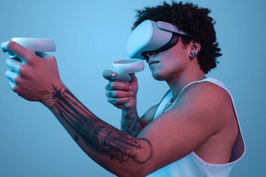 VR gözlüklü, kıvırcık saçlı ve dövmeli isimsiz bir erkek video oyunu oynuyor ve mavi arka planda sanal gerçeklik yaşıyor.