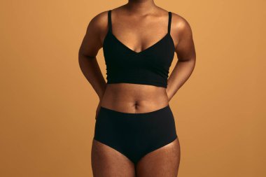 Siyah iç çamaşırlı Afro-Amerikan kadın model bej arka planda duruyor ve elleri arkada stüdyoda.