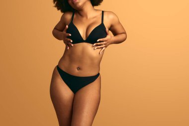 Göbeğinde piercing olan, siyah iç çamaşırı olan, turuncu arka planda dururken göğsüne dokunan isimsiz bir Afro-Amerikan kadın model.