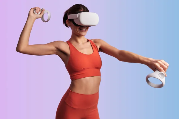 stock image Cheerful young female athlete with dark hair in sportswear and VR goggles playing virtual tennis with gamepads and smiling against two color background