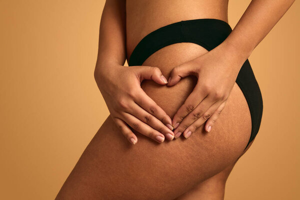 Анонимные урожай плюс размер этнической женщины в черных трусиках показывая жест любви на ягодицах на оранжевом фоне