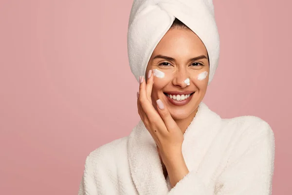 在粉红背景的护肤过程中 快乐的年轻女性穿着浴衣 头戴毛巾 在脸颊和鼻子上涂润肤霜 — 图库照片