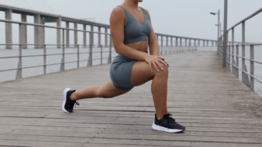 Genç bayan koşucu spor ve spor öncesi esneme hareketleri yapıyor. Gri spor giysili bir kadın dışarıda bacaklarını ısıtıyor..