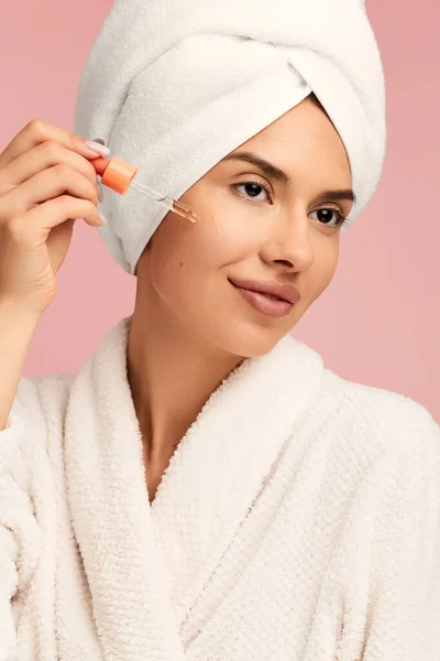 若い女性は ピンクの背景に対するスキンケア手順中に頬にピペットから血清を適用する頭の上にタオルで白いバスローブで — ストック写真