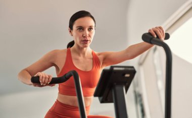 Düşük motivasyonlu, turuncu aktif giysili genç bir kadının spor salonundaki yoğun antrenman sırasında eliptik antrenör üzerinde egzersiz yapması.