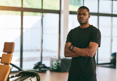 Ciddi kaslı Afrikalı Amerikalı erkek eğitmen modern fitness merkezinde kollarını kavuşturup kameraya bakıyor.