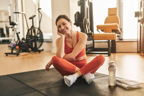 Full body of young positive athletic woman in orange activewear sitting on mat near bottle of water and towel, leaning on hand looking at camera after fitness training