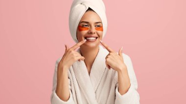 Göz bantlı, kafasında bornoz ve havlu olan iyimser genç bir bayan pembe arka planda dişlek bir gülümsemeyi işaret ediyor.