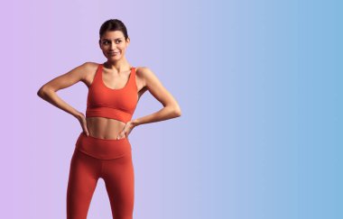 Genç bayan, kırmızı dar spor giyer, elleri belinde, arka planda durur ve başka tarafa bakar.