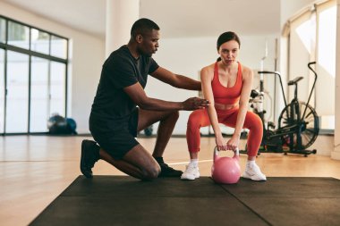 Full body of strong African American male instructor helping female athlete doing squats with pink kettlebell in gym during workout clipart