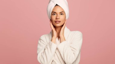 Beyaz bornoz ve havlu giymiş genç bir kadın pembe arka planda cilt üstüne yüz teması yapıyor.