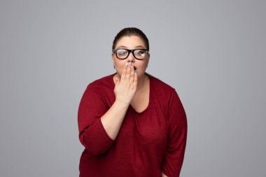 Hayretler içinde, koyu renk saçlı, gözlüklü, ağzı kapalı, elleri kapalı, gri arka planda kameraya bakan iri yarı, genç bir kadın.