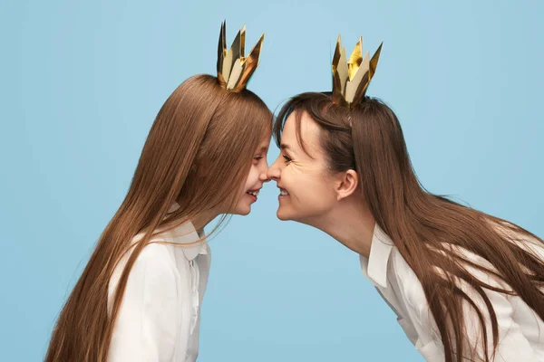 Side view of little girl and woman wearing golden crowns and making grimace to each other standing opposite on blue background