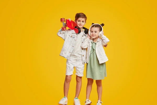 一对可爱的时尚孩子与滑板微笑和听音乐在耳机 而站在明亮的黄色背景 兄弟姐妹拥抱 — 图库照片