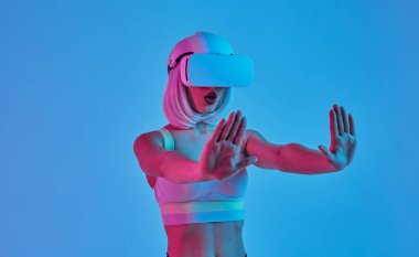 Fütüristik Vr gözlüklü şaşkın kadın film izlerken ve neon ışık altında mavi arka plan karşı dururken her iki eliyle stop jest gösteren