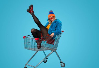 Moda kıyafetli, gülümseyen ve mavi arka planda alışveriş arabasında oturmuş kameraya bakan genç bir kadın.