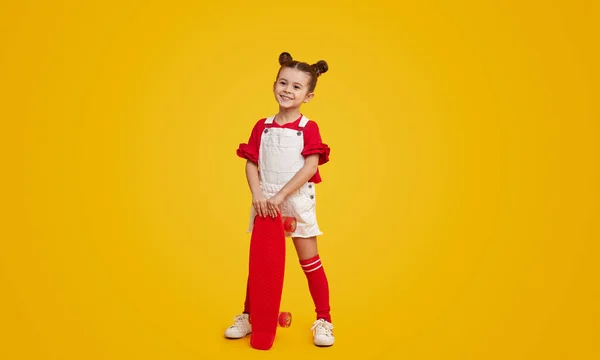 黄色の背景に明るい赤いロングボードでポーズをとりながらカメラを見てスタイリッシュな衣装でかわいい子供モデル — ストック写真