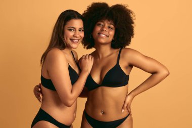 Sevinçli genç İspanyol ve Afro-Amerikan kadın artı siyah iç çamaşırlı koyu renk saçlı mankenler bej arka planda kameraya sarılıp gülümsüyorlar.