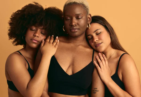 Tranquil multiracial female models in black lingerie embracing each other and closing eyes while standing together on beige background