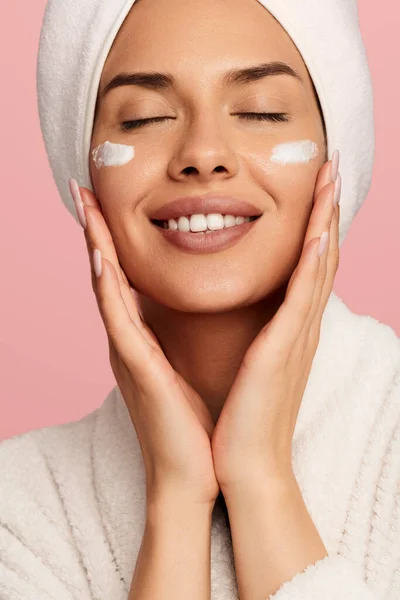 ピンクの背景に目を閉じた肌の美しさと優しさを楽しむ顔に保湿クリームを適用する頭にタオルが付いている幸せな若い女性 — ストック写真