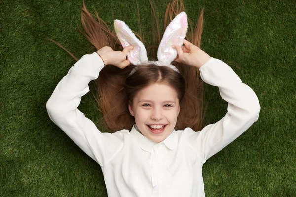上面的可爱的女孩在白色的耳朵摆姿势作为快乐的兔子 而躺在绿色的草地上 对着镜头微笑 — 图库照片