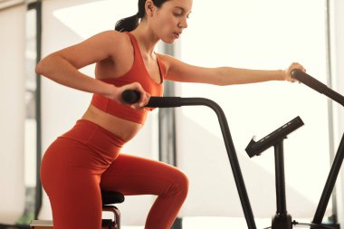 Kırmızı spor kıyafetli güçlü bayan sporcunun spor salonunda kardiyo antrenmanı sırasında eliptik makine üzerinde egzersiz yapması.