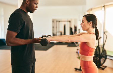 Spor salonunda egzersiz yapan kadın sporculara yardımcı olan güçlü erkek eğitmenin yan görüntüsü eğitim sırasında kollarını ileri doğru uzatarak ağır dambılları kaldırması.