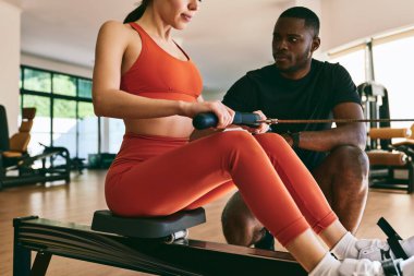 Ekin formalı bayan sporcu spor salonunda siyah kişisel eğitmenle antrenman yaparken kürek çekme makinesinde direnç bandıyla egzersiz yapıyor.