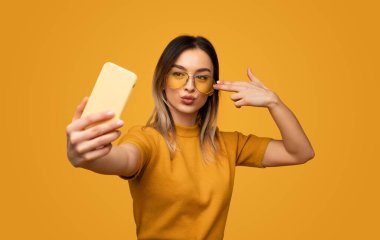 Parlak güneş gözlüklü şık genç kadın sarı arka planda selfie çekerken akıllı telefon arıyor.
