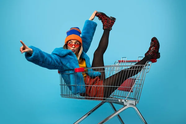 Full body of crazy young female in warm clothes sitting in shopping trolley while looking away against blue background