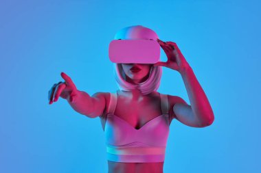 VR gözlüklü genç fütürist kadın neon ışıkta mavi arka planla sanal gerçeklikle etkileşime geçiyor