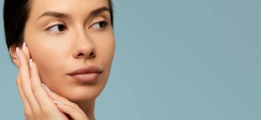 Genç bir kadın yüzlerine dokunuyor ve stüdyoda mavi arka plana bakıyor. Güzellik ve cilt bakımı