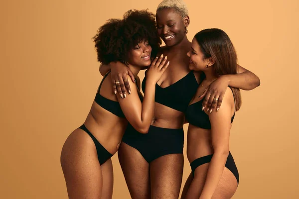 Group of multiethnic women in black lingerie embracing with eyes clothes while standing against beige background