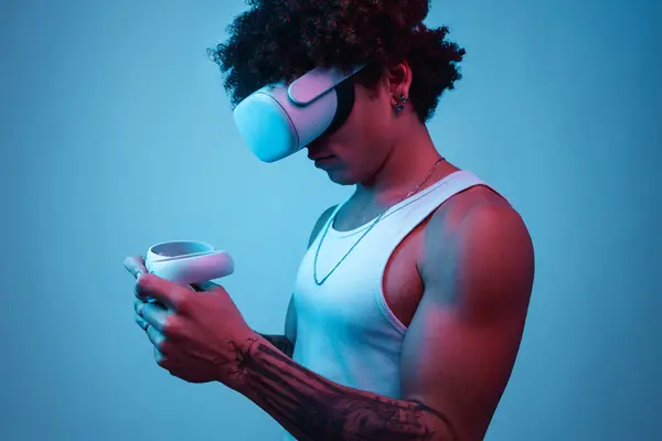 Vrゴーグルに立っている間 手元にタトゥーを持つ若いヒスパニック男性の側面ビュー 青い背景とのビデオゲームをプレイ中にコントローラをチェック — ストック写真