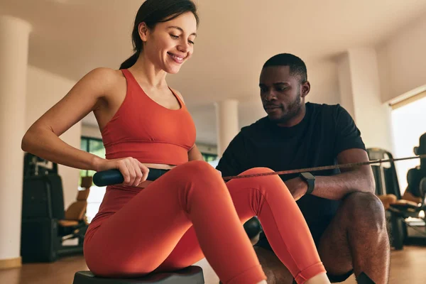 Strong positive female athlete in orange activewear doing exercise with strap on rowing machine during fitness training with black coach in gym