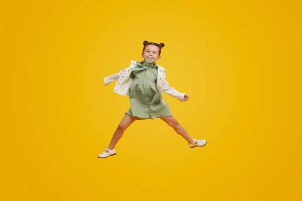 明るい黄色の背景にジャンプしながら スタイリッシュなドレススイング腕と笑顔でフルボディかわいい子供 — ストック写真