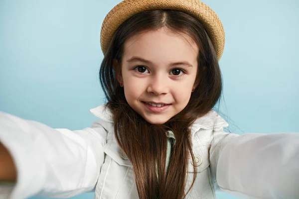 スタイリッシュなジャケットと帽子の小さなかわいい女の子は 青い背景に微笑んでいる間 セルフィーショットを撮ります — ストック写真