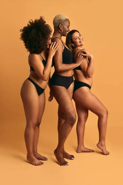 Full body side view of African American female models in lingerie standing against beige background in studio