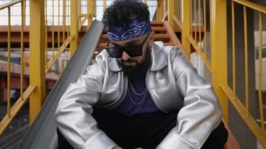 Dinamik bir yakınlaştırma sahnesinde kendine güvenen bir hip-hop dansçısı şehir ortamında yer alır. Parlak bir ceket, güneş gözlüğü ve saç bandı takmış, tutum ve yağma kavramı, modern şehir kültürünün somut bir örneği.