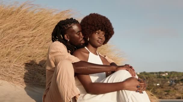 一对时髦的非洲黑人夫妇 在浪漫的时刻 在日落时坐在沙滩上的沙丘上 黑人男人拥抱着黑人非洲女人 窃窃私语 分享欢笑 深深地坠入爱河 — 图库视频影像