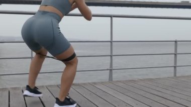 Spor kıyafetleri içinde tanınmayan fit bir kadın dışarıda antrenman yapıyor, kalça kaslarını hedef almak için lastik bantla yan adımlar atıyor.