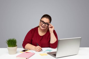 Pozitif genç, iri yarı, siyah saçlı, günlük giysiler içinde, gözlük takan ve kameraya gülümseyen, masa başındaki dizüstü bilgisayarda çalışırken not alan bağımsız bir kadın.