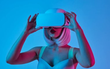 Şehvetli genç bayan sütyen ve pembe peruk Vr gözlük giyen ve mavi arka plan karşı neon ışık altında dururken etrafa bakıyor