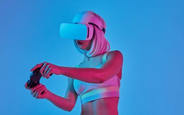 Mavi arka plan karşı neon ışık altında dururken video oyunu oynamak için denetleyici kullanarak modern Vr kulaklık fütüristik genç kadın