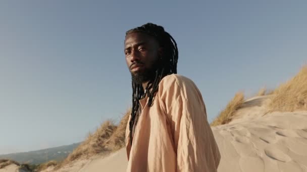 スタイリッシュな夢とゆったりとしたオープンシャツを持つアフリカの黒人男性は 砂の砂丘の背景にカメラに回しながら自信を誇ります — ストック動画