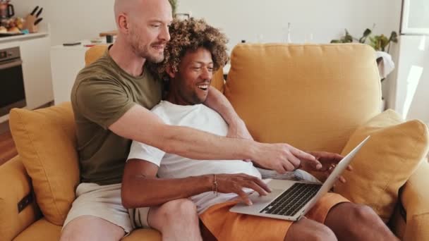 一对多样化的同性恋夫妇 一个黑发男人和他的白人伴侣 兴高采烈地浏览了一下网络 预订了度假票 并在家里的沙发上分享了旅行计划 — 图库视频影像
