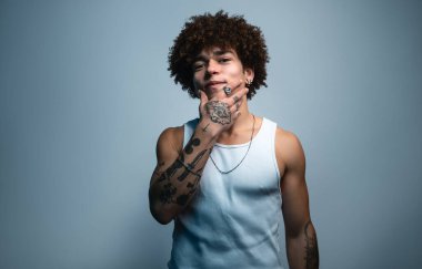 Kıvırcık saçlı, kolları dövmeli, çenesine dokunan ve kameraya bakan, kendine güveni tam İspanyol bir erkek.