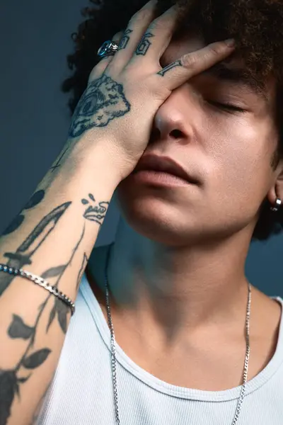 Closeup artistic Portrait of young ethnic male with with multiple tattoos on hand posing and covering eye with hand with eyes closed