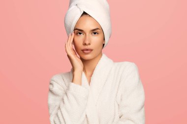 Kafasında havluyla tapınağa dokunan güzel bir kadın pembe arka planda kameraya bakarak hijyenik rutinden sonra stüdyoda dikilirken.