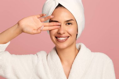 Bornozlu ve kafasında havlu olan mutlu genç bir kadın pembe arka planda kameraya bakarken gözünü silah jesti ile kapatıyor.